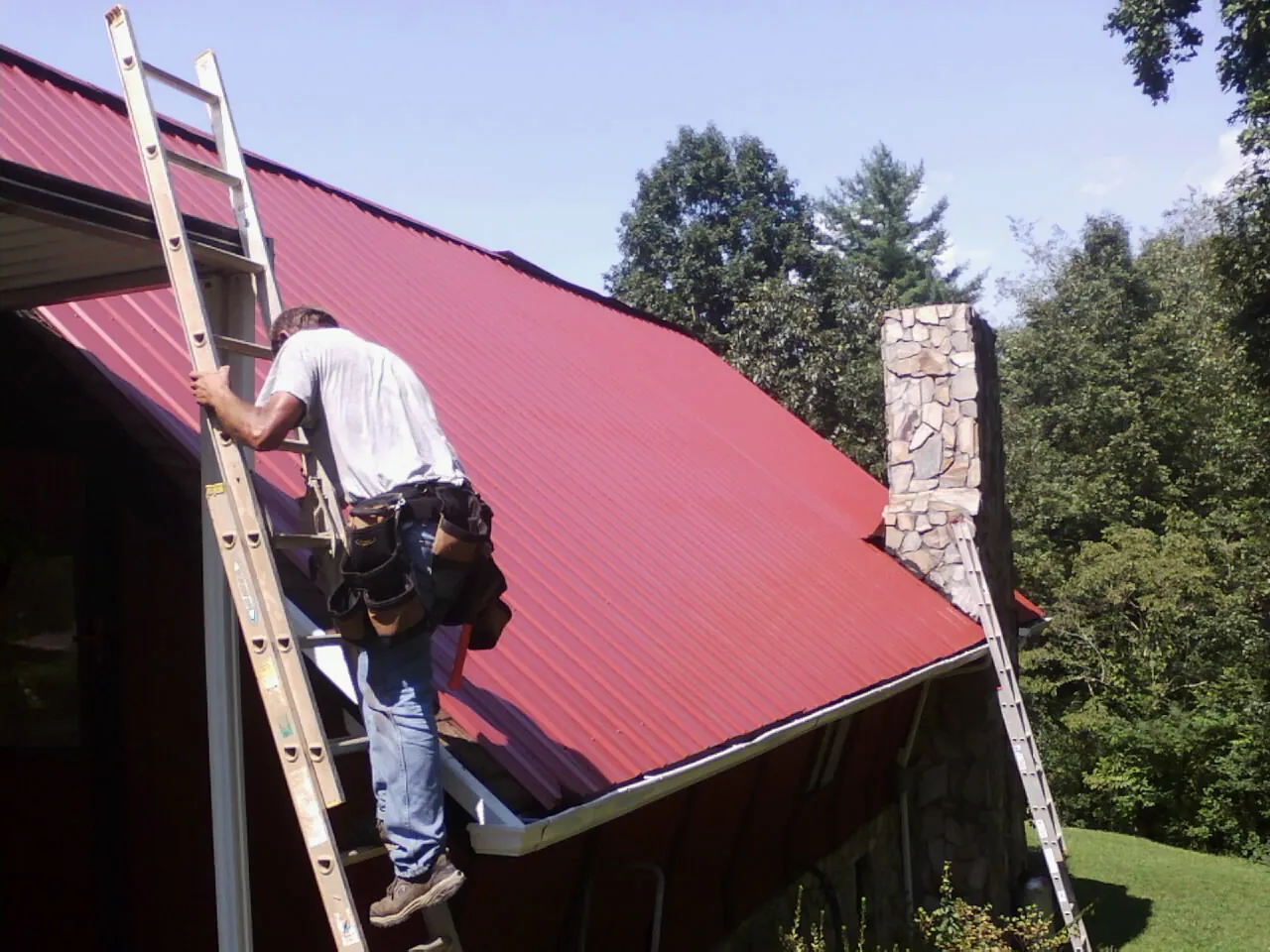 Roofer on a ladder beside a red metal roof and stone chimney — Soffit & Fascia Repair by Roofing Brothers in Tampico